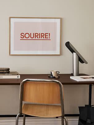Skrivbord med stol, lampa och inramad poster med texten "SOURIRE!" på väggen