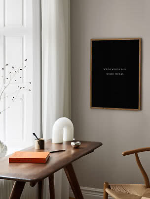Schwarzes Poster mit Zitat im Holzrahmen über einem Schreibtisch am Fenster in minimalistischem Raum