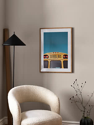 Framed poster of a yellow school bus on blue background above a white armchair beside a black floor lamp