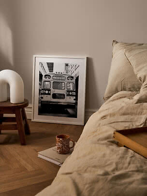 Chambre cosy avec linge de lit beige, tabouret en bois, lampe blanche et affiche de bus en noir et blanc