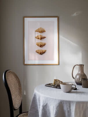 Gerahmtes Poster mit vier Croissants über einem Tisch mit Tasse, Krug und Kuchen.