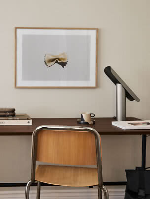 Minimalistisk skrivebord med træstol, lampe, kop og indrammet pastaplakat på væggen
