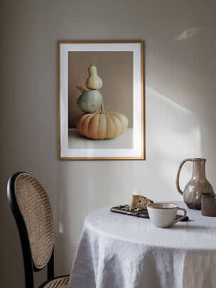 Framed poster of stacked pumpkins on a beige background above a table set with cup and pitcher.