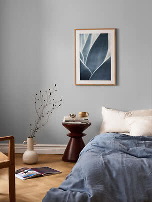 Schlafzimmer mit blauer Bettdecke, weißen Kissen und gerahmtem botanischem Bild an grauer Wand