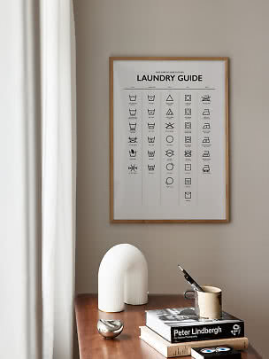 Gerahmtes Wäscheguide-Poster über einem Holztisch mit Büchern, Tasse und weißer Deko