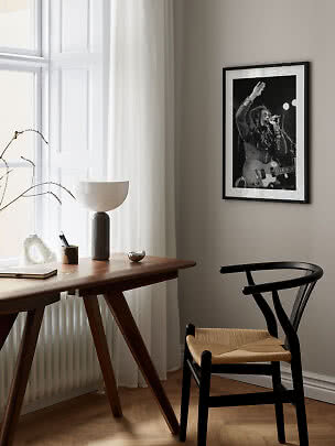 Indrammet sort-hvid koncertplakat på beige væg over skrivebord og sort stol