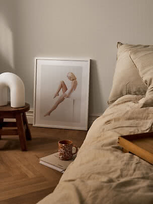 Chambre cosy avec linge de lit beige, affiche encadrée, lampe blanche et mug au sol