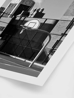 Black-and-white photo of a tilted basketball hoop by a fenced urban court