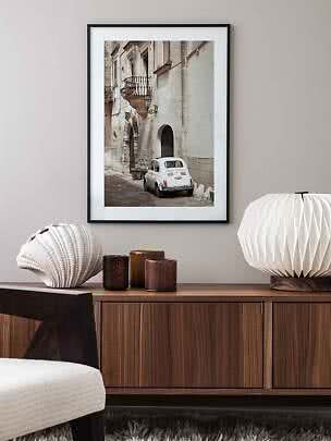 Framed print of a white vintage car on a narrow old street above a wooden sideboard