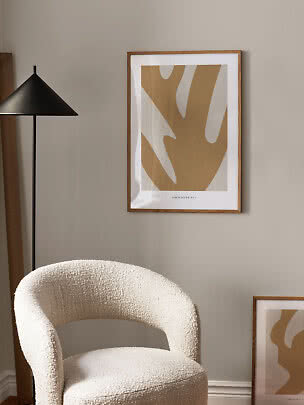 Abstracte beige poster in lichte houten lijst naast een zwarte vloerlamp en een witte fauteuil