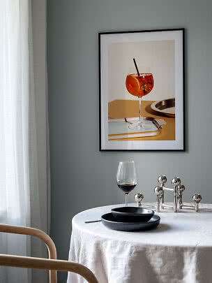 Eethoek met ronde tafel, zwart servies, wijnglas en ingelijste drankposter aan grijze muur