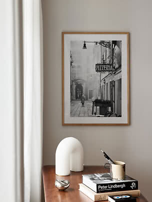 Framed black-and-white photo print of a narrow street with pizzeria sign on a beige wall