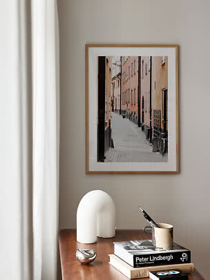 Framed photo of a narrow cobblestone street above a wooden table with books, vase and mug