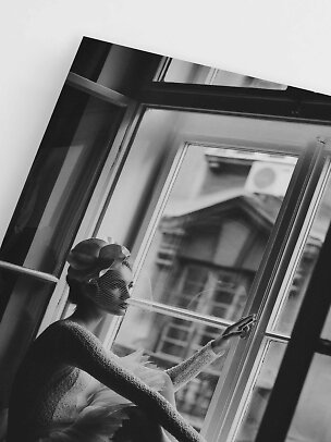 Photo en noir et blanc d’une femme en robe de tulle avec voile et fleurs près d’une fenêtre ouverte