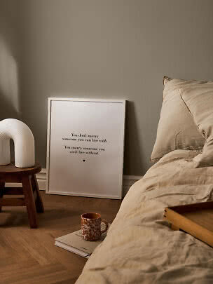 Schlafzimmer mit beiger Bettwäsche, gerahmtem Spruchposter, Holzschemel mit weißer Lampe und Tasse.
