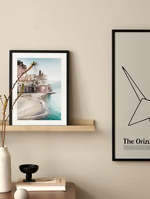 Black-framed coastal print on a wooden shelf above a table with vase, book and black bowl