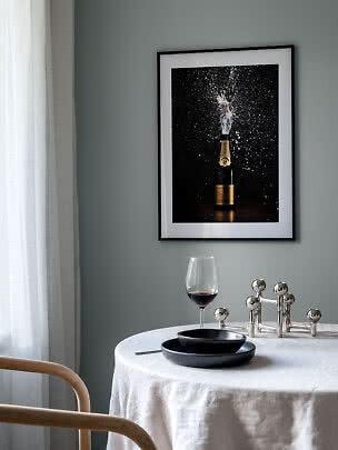 Póster enmarcado de una botella de champán estallando sobre una mesa puesta con copa y platos negros