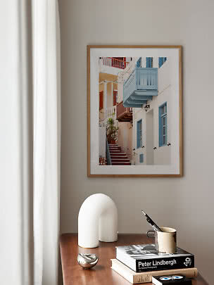 Framed photo print of a narrow street with stairs and blue balconies on a beige wall above a wood table