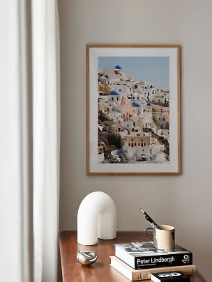 Framed photo print of a white coastal town with blue domes above a wooden table with books and decor