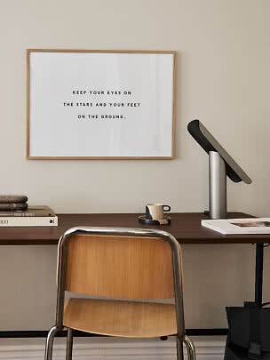 Skrivebord med træstol, lampe, kop og indrammet citatplakat på væggen.