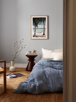 Schlafzimmer mit blauer Bettwäsche und gerahmtem Strandbild mit Hängematte an grauer Wand