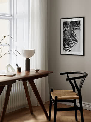 Black framed palm leaf print hanging above a wooden desk by a bright window.