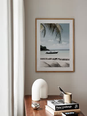 Lámina enmarcada de playa con barco y palmera sobre una mesa de madera con libros y decoración