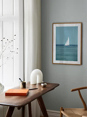 Framed sailboat print on a gray wall above a wooden desk in a calm modern interior