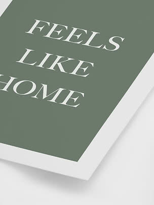 Green poster with white 'FEELS LIKE HOME' text in a white frame, shown at an angle