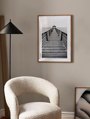 Framed black-and-white photo of a wooden pier over water on a beige wall beside a chair and black floor lamp
