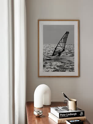 Ingelijste zwart-wit poster van een windsurfer boven een houten tafel met boeken en decoratie
