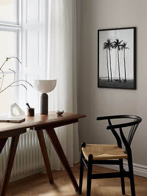 Schwarz gerahmter Palmenprint über Holzschreibtisch und schwarzem Stuhl am hellen Fenster