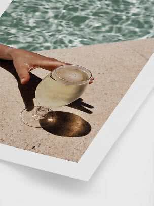 Hand greift nach einem Glas mit hellem Getränk am sonnigen Poolrand