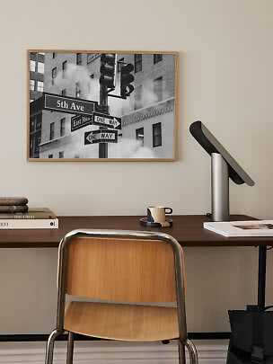 Skrivebord med stol og lampe under indrammet sort-hvid plakat af gadeskilte på 5th Ave.