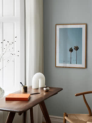 Framed palm tree print on a light blue background above a wooden desk by a window.