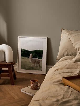 Gemütliches Schlafzimmer mit beiger Bettwäsche, Holzschemel, weißer Lampe und gerahmtem Alpaka-Poster