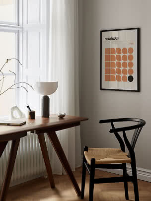 Bauhaus-plakat i sort ramme over træskrivebord med lampe og sort stol ved vindue