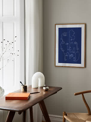 Affiche encadrée bleue à visages abstraits au-dessus d’une table en bois dans une pièce lumineuse