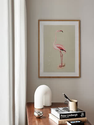 Framed flamingo print on a beige wall above a wooden table with books and decorative objects