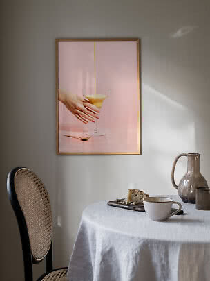 Inramad konstposter över matbord med rosa motiv av hand, glas och upphälld dryck