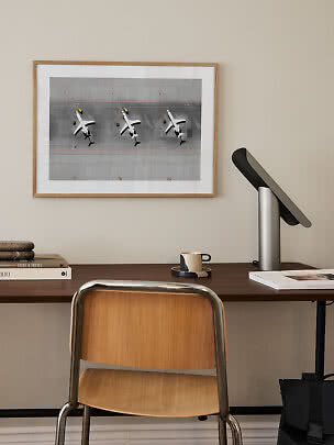 Indrammet plakat med fly set ovenfra hænger over et skrivebord med lampe og stol