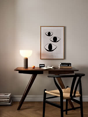 Træskrivebord med sort stol, tændt bordlampe og indrammet kunstplakat med tre stiliserede øjne.