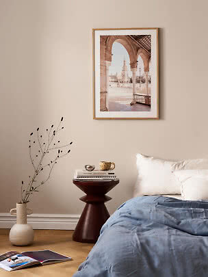 Soveværelse med seng, rundt sidebord og indrammet arkitekturplakat på beige væg