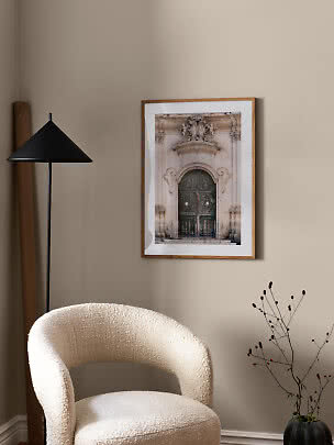 Póster enmarcado de una puerta ornamentada en pared beige sobre una silla crema texturizada.