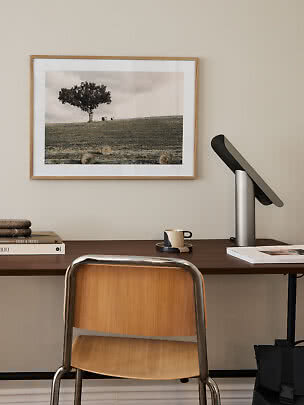 Skrivebord med stol, sort bordlampe og indrammet landskabsplakat på væggen