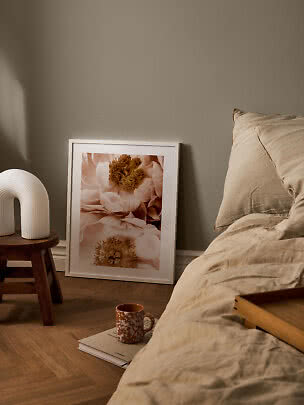 Schlafzimmer mit beiger Bettwäsche, Blumenbild im weißen Rahmen und weiße Lampe auf Holzschemel