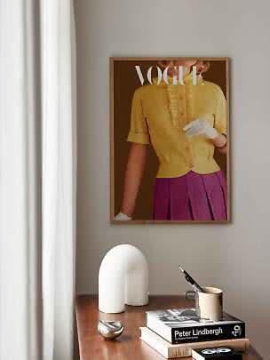 Gerahmtes Vogue-Poster an der Wand über Holztisch mit Büchern, Tasse und weißer Dekoleuchte
