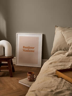 Gemütliches Schlafzimmer mit beiger Bettwäsche, gerahmtem Bonjour Madame-Poster und weißer Lampe