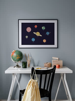 Oprawiony plakat z planetami nad białym biurkiem z globusem, pędzlami i czarnym krzesłem.