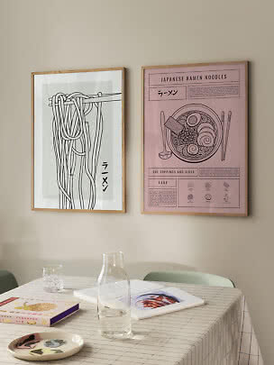 To indrammede ramen-plakater på beige væg over et bord med glasflaske, bøger og tallerken